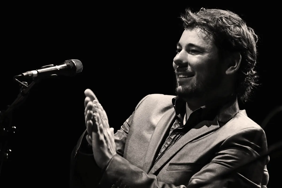 A flamenco singer smiling and clapping palmas in front of a microphone on a dark stage